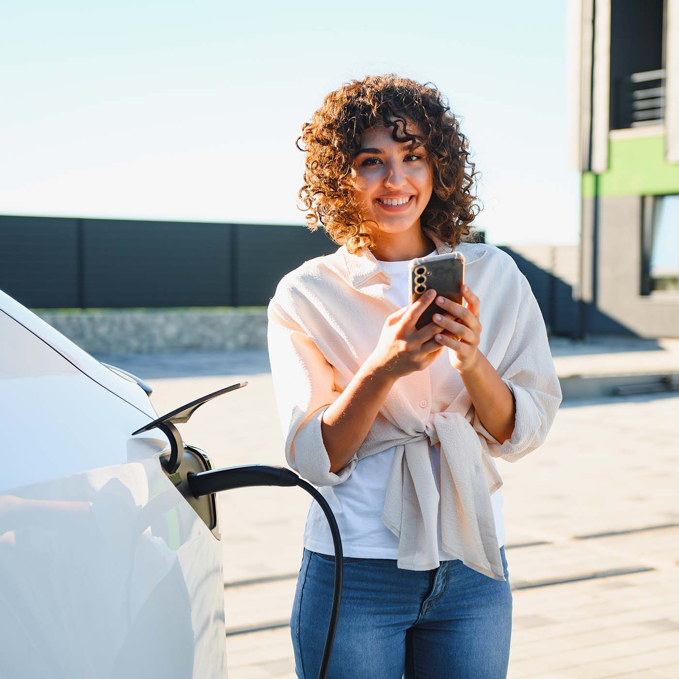 Happy woman using smartphone while charging an electric car in front of her house. Happy woman using smartphone while charging an electric car in front of her house.
